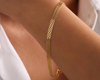 Op de afbeelding: Een goudkleurige armband met een geweven ontwerp wordt getoond op een arm. De armband is gemaakt van kleine, rechthoekige schakels, waardoor een textuur ontstaat. De achtergrond omvat een wit overhemd.