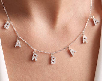 May include: A silver necklace spelling out the name "BARBARA" in individual, sparkling letter charms. The necklace hangs on a delicate chain, and the letters are evenly spaced. The background is a white top.