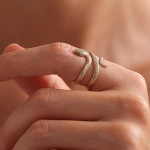May include: A gold-toned ring shaped like a coiled snake that wraps around the finger. The metal surface features a fine, textured scale pattern throughout the body, with a small, distinct head and a pointed tail end visible on the finger.