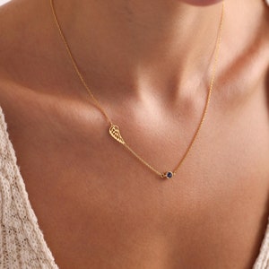May include: A gold chain necklace with a single wing pendant and a small blue gemstone. The necklace is worn by a person.