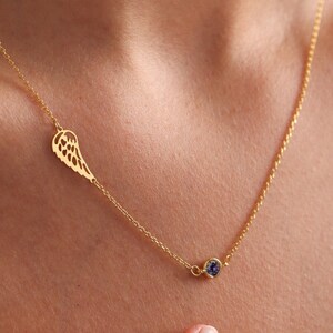May include: A gold chain necklace with a delicate wing pendant and a small blue gemstone. The necklace is worn by a person with their hand on their chest.