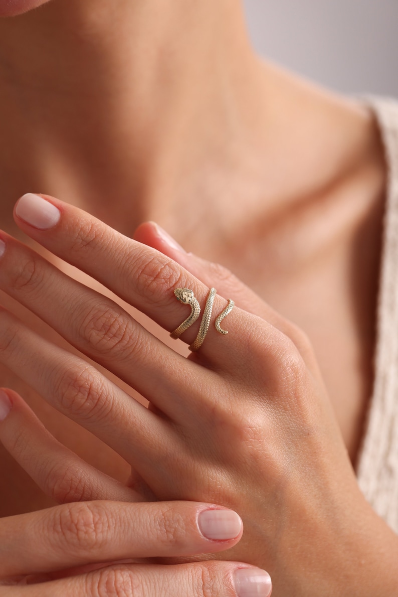 May include: A gold-colored snake ring. The ring wraps around the finger, with the snake's head and tail forming the ends. The snake's head has small details, and the ring is worn on a hand.