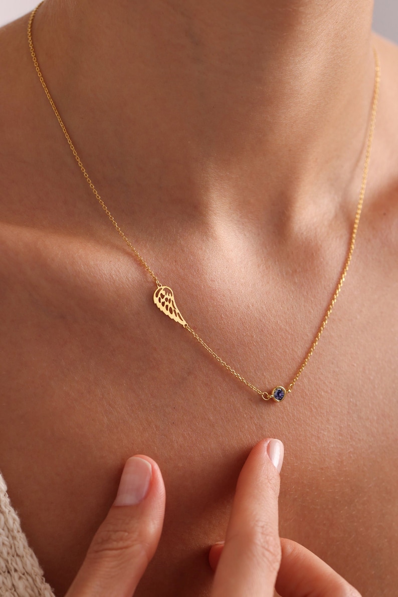 May include: A gold chain necklace with a delicate wing pendant and a small blue gemstone. The necklace is worn by a person with their hand on their chest.
