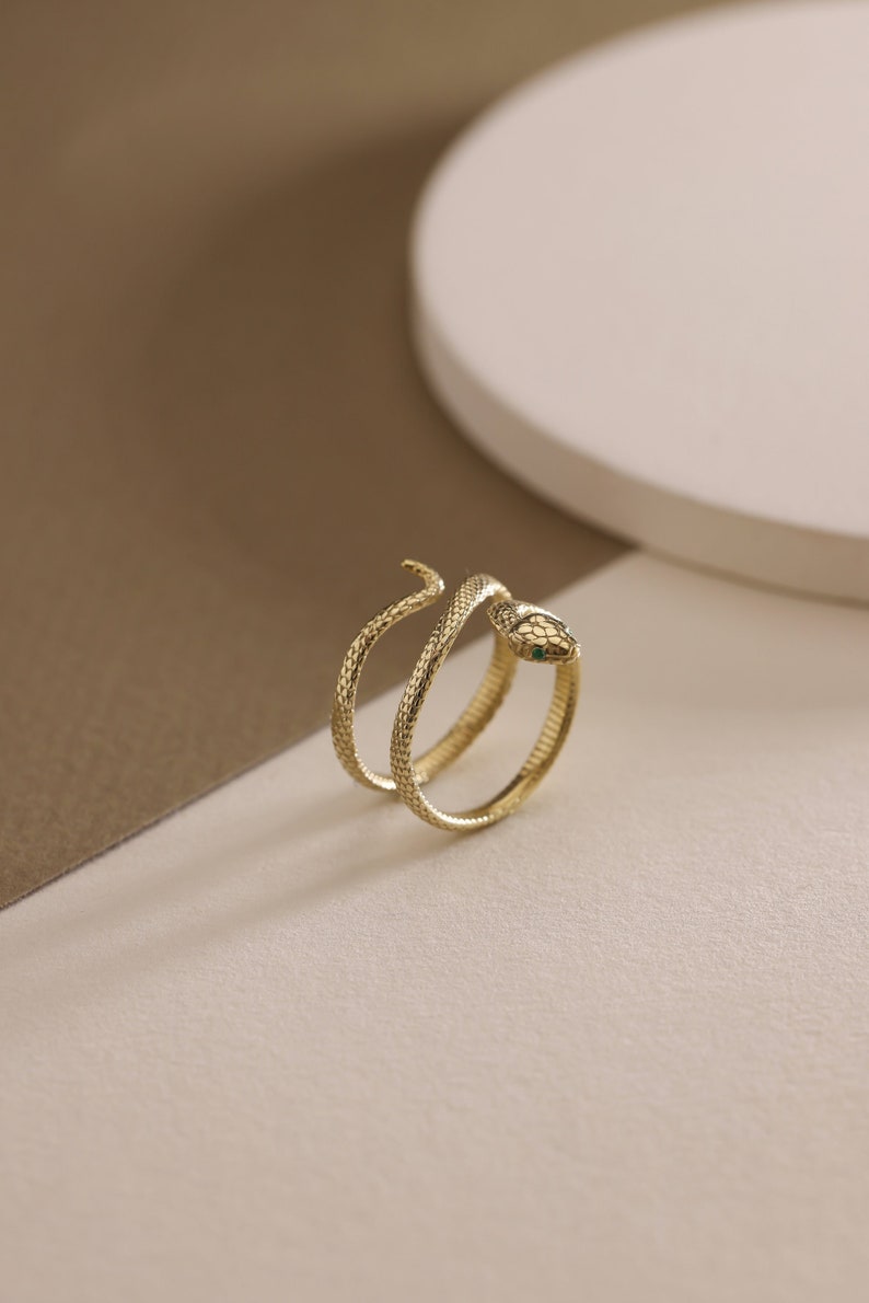 May include: A gold-colored snake ring with a textured, scale-like pattern. The snake's head features a small green stone. The ring wraps around the finger in a spiral design.
