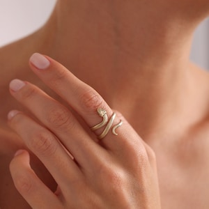 May include: A gold-colored snake ring with a textured design. The ring wraps around the finger, with the snake's head and tail forming the ends. The ring is worn on a person's finger.