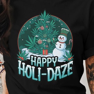 Könnte beinhalten: Schwarzes T-Shirt mit festlichem Design. Die Grafik zeigt einen Weihnachtsbaum aus Cannabisblättern, einen Schneemann und die Worte "Happy Holi-Daze" in einer winterlichen Schriftart. Das Design umfasst rote, grüne, weiße und blaue Farben.