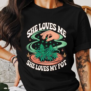 Puede incluir: Camiseta negra con un diseño gráfico blanco de una mano sosteniendo plantas de marihuana. El texto "She loves me, She loves my pot" está impreso en la camiseta.
