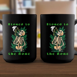 May include: Two black coffee mugs with a skeleton smoking a joint and surrounded by marijuana leaves. The text on the mugs reads "Stoned to the Bone".