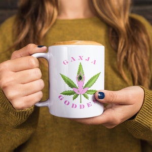 May include: White ceramic mug with a green marijuana leaf design and the text "GANJA GODDESS" in pink letters. A pink silhouette of a person in a yoga pose is inside the leaf.