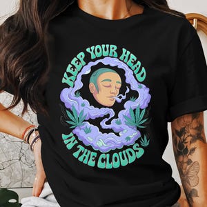 May include: Black t-shirt with a graphic design of a person with green hair and a green plant in a cloud of smoke. The text "Keep Your Head In The Clouds" is written in a retro font.
