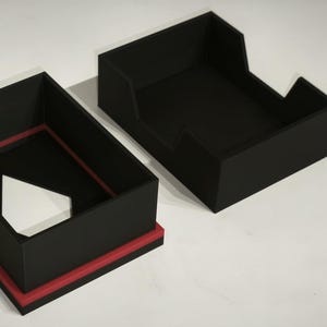 Peut inclure: Boîte noire en deux parties avec un accent rouge. La boîte est ouverte, révélant une forme triangulaire blanche à l'intérieur. La deuxième pièce est une structure en forme de plateau. La boîte est sur une surface blanche.