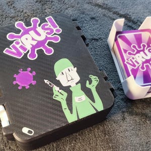 Könnte beinhalten: Eine schwarze Kartenspielbox und ein Kartenhalter mit dem Wort "VIRUS!" in Lila. Die Box hat eine Grafik einer Person in Grün, die eine Spritze hält. Der Kartenhalter enthält zwei Kartenspiele mit dem gleichen "VIRUS!"-Design.