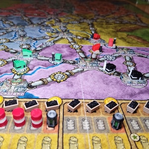 Puede incluir: Un tablero de juego con un mapa morado y verde que presenta las ciudades de Constanza, Augsburgo y Múnich. Hay pequeñas piezas de juego rojas, verdes y negras en el tablero. También hay pequeñas cajas de madera con piezas negras en su interior y barriles rojos.