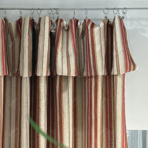Peut inclure: Rideaux de fenêtre à rayures rouges, beiges et vertes. Les rideaux sont suspendus à une tringle argentée avec des anneaux métalliques. Le tissu présente des rayures verticales et une cantonnière à volants en haut.