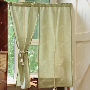 May include: Light green linen curtains with a neutral-colored tie and tassels. The curtains are hanging in front of a window, with a wooden cabinet and chair visible in the background. The word "WOGOL" is printed on the curtains.