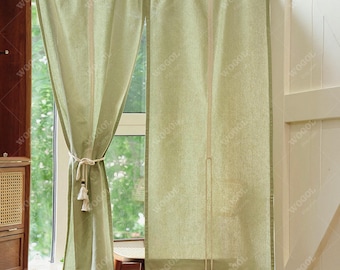 May include: Light green linen curtains with a neutral-colored tie and tassels. The curtains are hanging in front of a window, with a wooden cabinet and chair visible in the background. The word "WOGOL" is printed on the curtains.