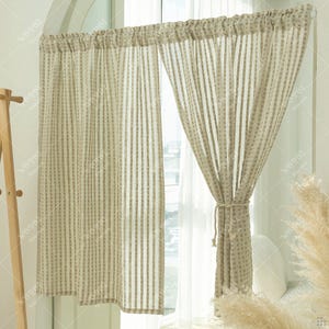 Puede incluir: Cortinas transparentes a rayas beige y blancas colgadas frente a una ventana. Las cortinas están atadas con una cuerda. La tela tiene un patrón texturizado y punteado. Tema de decoración del hogar.