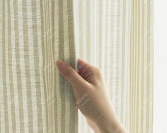 Puede incluir: Panel de cortina a rayas beige y blancas. Las rayas verticales crean un diseño clásico y neutro. La tela parece ser un material ligero, similar al lino. Una mano sostiene la cortina, oscureciendo parcialmente la vista.