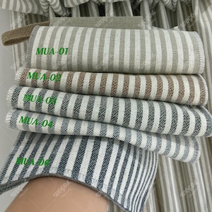 Op de afbeelding: Een stapel gestreepte stofstalen in verschillende kleuren, waaronder beige, bruin en grijs. Elk staal is voorzien van een groene code. De stoffen zijn gevouwen en opgestapeld, waarbij de verschillende kleuropties en patronen worden getoond.