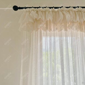 May include: Cream-colored sheer curtains with a ruffled valance. The curtains hang from a black rod, creating a soft, airy look. The valance adds a touch of elegance to the window treatment. The curtains filter light, creating a warm and inviting atmosphere.