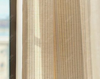 May include: A light beige curtain panel featuring a series of thin, vertical dark brown pinstripes. The fabric has a visible woven texture and a semi-sheer quality, allowing light to pass through the material while hanging in soft folds.