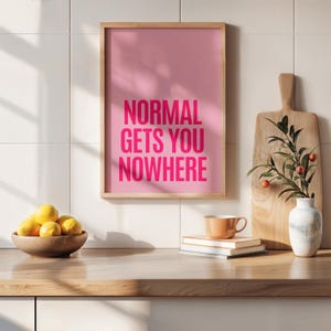 May include: A framed pink print with the words "NORMAL GETS YOU NOWHERE" in bright pink. The print is displayed on a wooden countertop with a bowl of lemons, a cutting board, and a small cup.