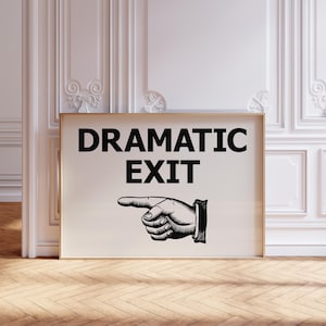 May include: A framed print with the words "DRAMATIC EXIT" in bold black letters. Below the text is a black and white illustration of a hand pointing to the right. The print has a gold frame and is displayed against a white wall.
