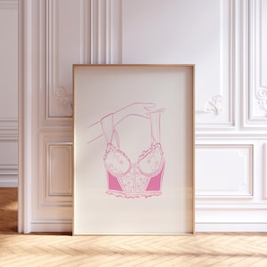 Op de afbeelding: Een ingelijste print met een roze lijntekening van een korset dat door een hand wordt vastgehouden. Het korset heeft bloemendetails en een gerimpelde rand. De achtergrond is lichtbeige en de lijst is lichtbruin.