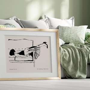 Puede incluir: Una impresión artística enmarcada con una ilustración minimalista en blanco y negro de una persona reclinada en una silla. La impresión está en un marco de madera clara y está etiquetada como "Poster No 01" y "Art Gallery, New York". La obra de arte se exhibe en una cama con almohadas y una manta verde.