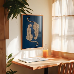 May include: A framed art print featuring two cheetahs with black spots on a dark blue background. The cheetahs are facing each other. The frame is a light brown color. The print is hanging on a white wall.