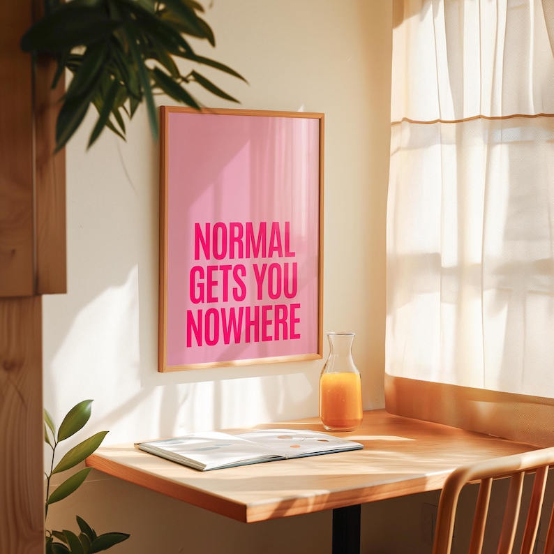 May include: A framed pink poster with the text "NORMAL GETS YOU NOWHERE" in bold, fuchsia letters. A glass carafe of orange juice sits on a wooden desk with an open book. The scene is lit by sunlight.