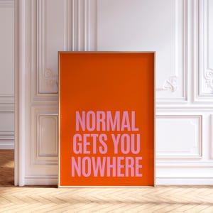 May include: An orange framed art print with the text "NORMAL GETS YOU NOWHERE" in pink. The artwork is displayed against a white wall with decorative molding and a wooden floor.