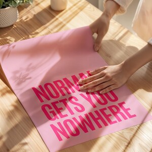 May include: A pink poster with the words "NORMAL GETS YOU NOWHERE" in bright pink block letters. The poster is being held by a person on a wooden surface. The poster is a motivational or inspirational item.