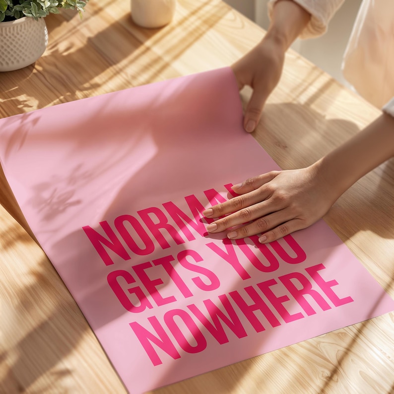 May include: A pink poster with the words "NORMAL GETS YOU NOWHERE" in bright pink block letters. The poster is being held by a person on a wooden surface. The poster is a motivational or inspirational item.