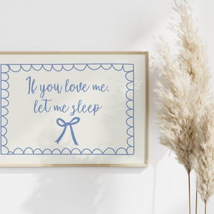 Puede incluir: Una impresión enmarcada con el texto "If you love me, let me sleep" en escritura azul, enmarcada por un borde festoneado azul. Un lazo azul está debajo del texto. La impresión se muestra junto a hierba ornamental alta y seca.