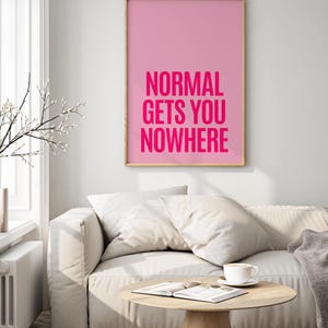 May include: A framed pink art print with the text "NORMAL GETS YOU NOWHERE" in bright pink. The artwork hangs above a white sofa with pillows, a wooden coffee table, and a cup of coffee.