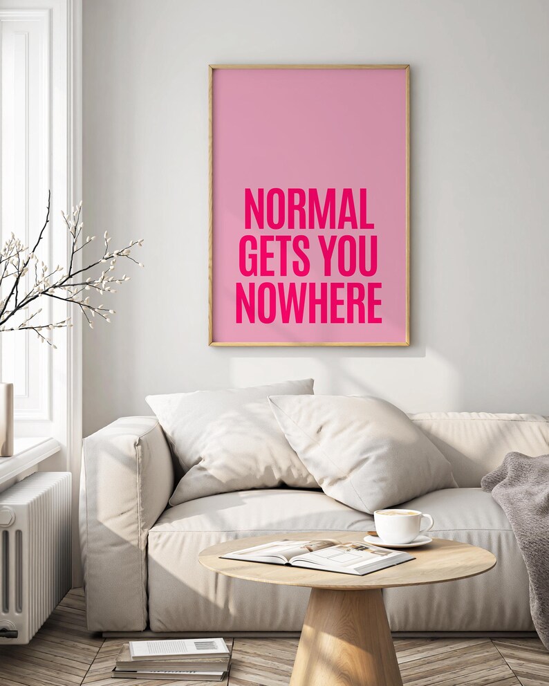 May include: A framed pink art print with the text "NORMAL GETS YOU NOWHERE" in bright pink. The artwork hangs above a white sofa with pillows, a wooden coffee table, and a cup of coffee.