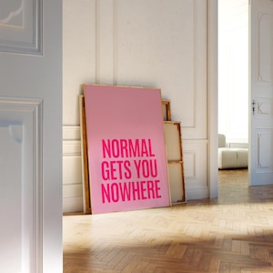 May include: A pink poster with the text "NORMAL GETS YOU NOWHERE" in a bold, fuchsia font. The poster is leaning against a white wall, with a wooden frame. The image is taken indoors, with a glimpse of a room in the background.