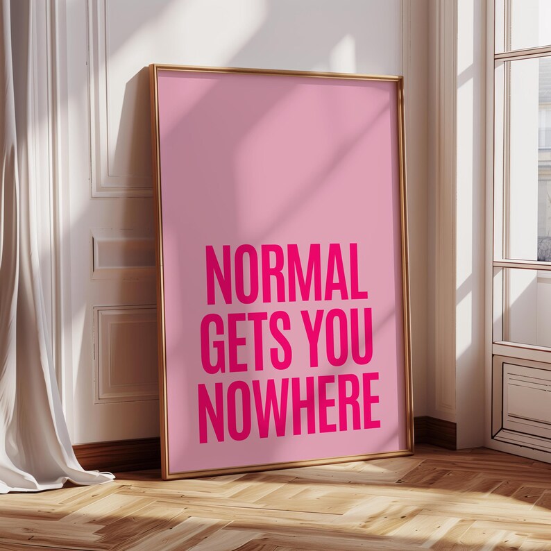 May include: A framed pink art print with the words "NORMAL GETS YOU NOWHERE" in bold, hot pink letters. The frame is gold, and the print leans against a white wall and wooden floor.