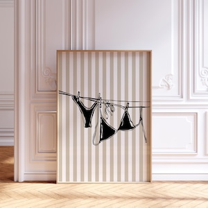 May include: A framed art print featuring a black and white line drawing of a bikini set hanging on a clothesline. The background has vertical beige and white stripes. The artwork is displayed in a room with a wooden floor and white walls.