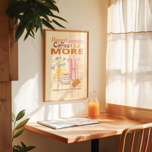 May include: A framed poster with the text "Happy Sunday Coffee Club MORE" and an illustration of a coffee pot, flowers, and a croissant. A glass carafe of orange juice and a book sit on a wooden table.