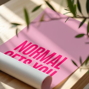 May include: A rolled-up pink poster with the words "NORMAL GETS YOU" in bright pink lettering. The poster is partially unrolled, revealing the text. The background is a light wooden surface with green leaves.
