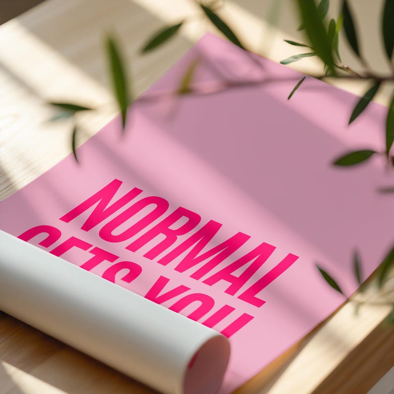 May include: A rolled-up pink poster with the words "NORMAL GETS YOU" in bright pink lettering. The poster is partially unrolled, revealing the text. The background is a light wooden surface with green leaves.