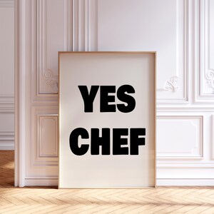 Op de afbeelding: Een ingelijste print met de woorden "YES CHEF" in vette zwarte letters op een crèmekleurige achtergrond. De print zit in een gouden lijst en staat tegen een witte muur met decoratieve lijstwerk en een houten vloer.