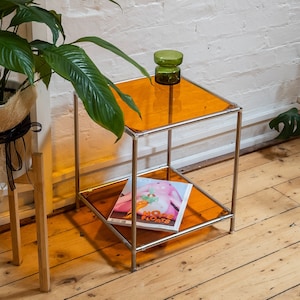 May include: A small, two-tiered side table with a chrome frame and amber glass shelves. A magazine with the text "Hope Floats" is on the lower shelf. A green glass vase sits on the top shelf.