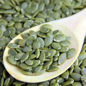 May include: A wooden spoon filled with a mound of raw, oval-shaped, dark green pumpkin seeds. The spoon is positioned over a larger pile of identical pumpkin seeds, showcasing their smooth texture and consistent deep green colour throughout.