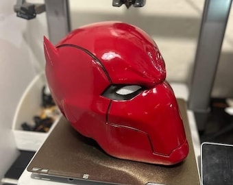 May include: A glossy red helmet with a distinctive design, featuring angular lines and eye openings. The helmet is placed on a printer bed, suggesting it's a 3D-printed item. The overall aesthetic hints at a superhero or cosplay theme.