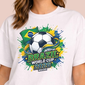 May include: White t-shirt featuring a soccer ball graphic with the text "BRAZIL WORLD CUP 2026 SOCCER". The design includes the Brazilian flag and colorful paint splatters in green, yellow, and blue.