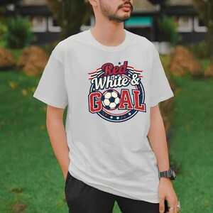 May include: White t-shirt with a soccer-themed graphic. The design features the text "Red White & Goal" in red and blue, with a soccer ball in the center. The shirt is a classic crew neck style.