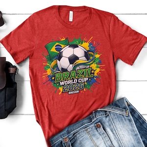 K&ouml;nnte beinhalten: Rotes T-Shirt mit einem Fu&szlig;ball-Grafik, der brasilianischen Flagge und dem Text "BRAZIL WORLD CUP 2026 SOCCER". Das Design umfasst farbenfrohe Farbspritzer in Gelb, Blau und Gr&uuml;n. Das Shirt wird mit einer blauen Jeans kombiniert.